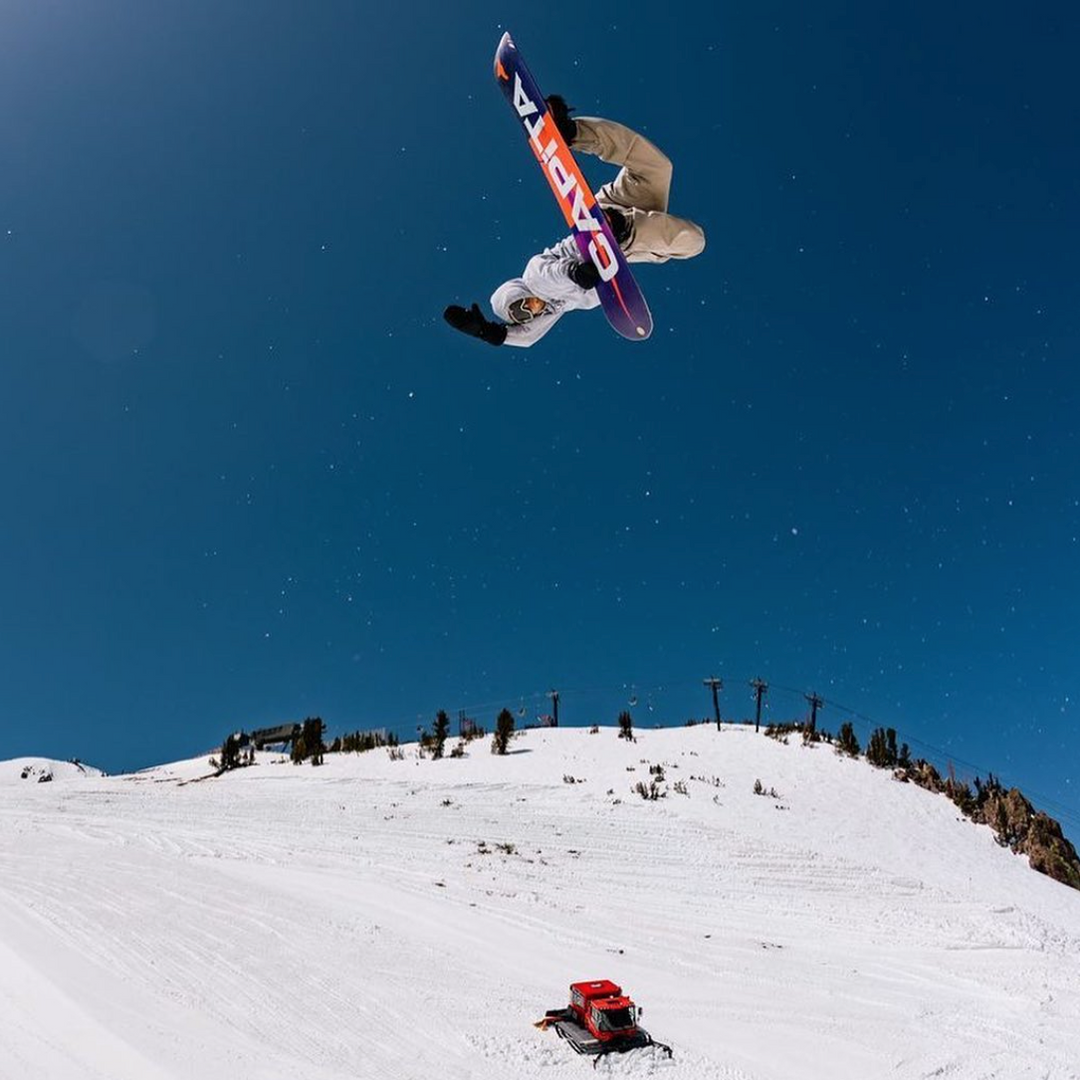 Brandon Cocard flipping upside down on a snowboard high above a groomed slope and snowcat