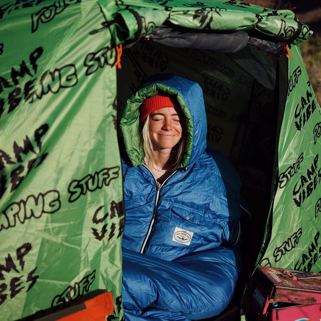 Britten Duerden smiling in a blue hooded sleeping bag inside a green "Camp Vibes" print tent