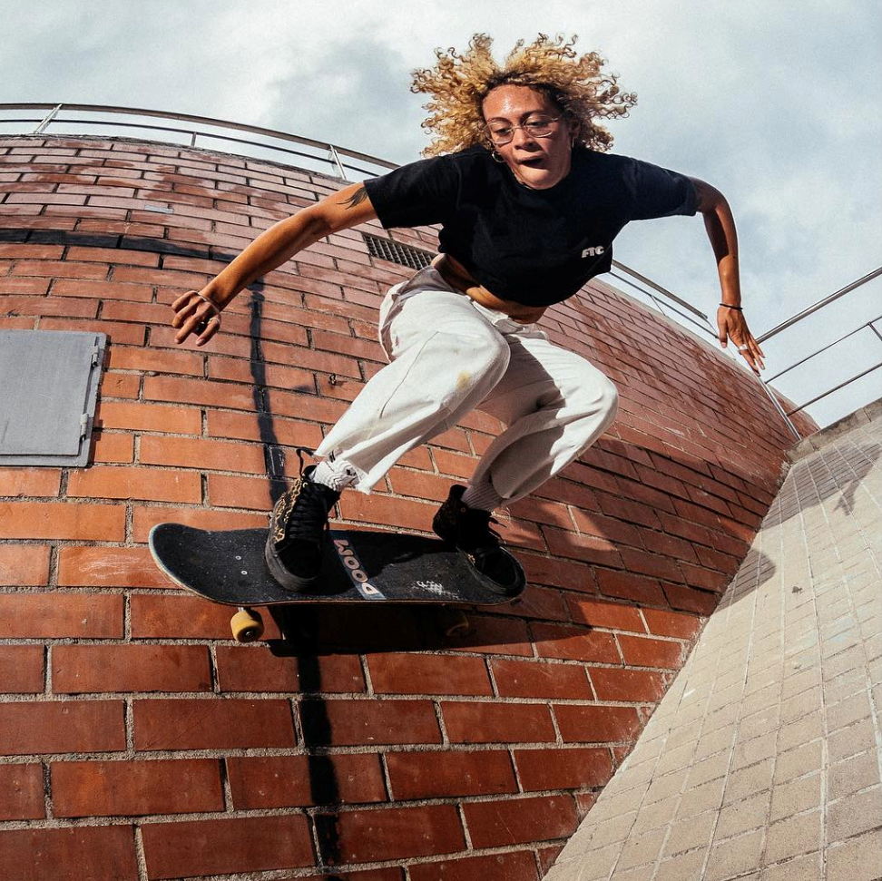 Dayana Young riding a skateboard up a curved red brick wall in a black tee and white pants