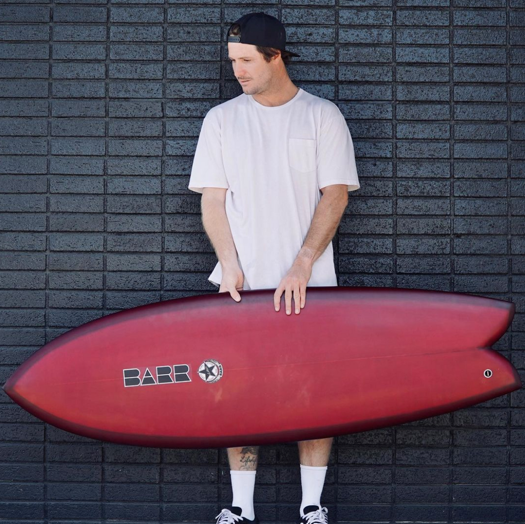 Duran Barr holding a red Barr fish surfboard in front of a dark brick wall
