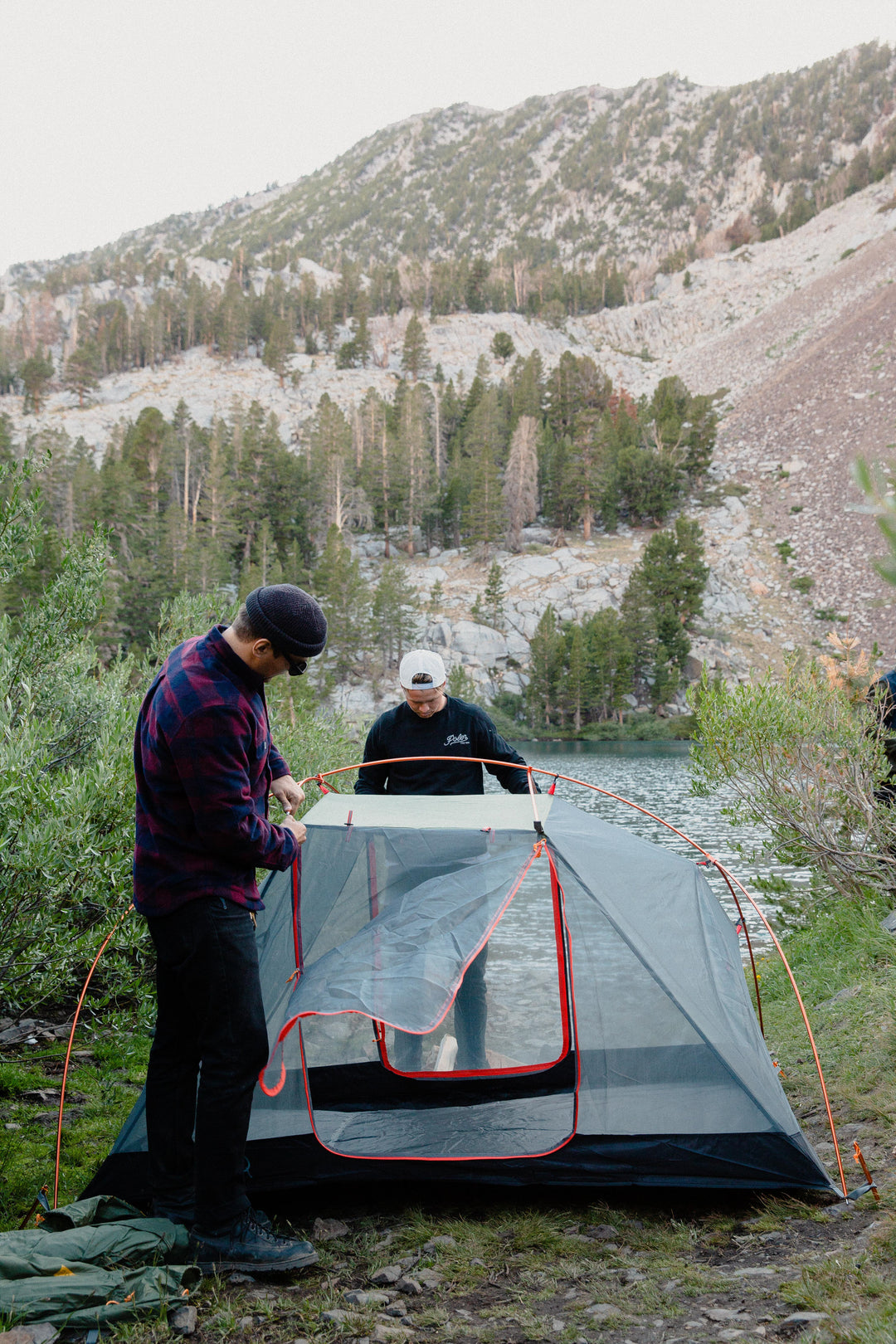 Two campers setting up a gray Poler tent with orange poles by a lake below a rocky, forested mountain