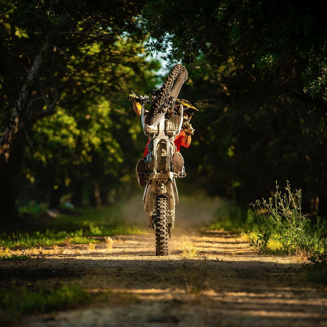 Jimmy Hill popping a wheelie on a dirt bike down a sunlit, tree-lined dirt road