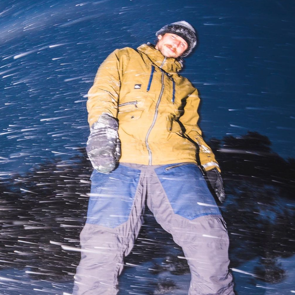 Justus Hines in a mustard snow jacket and blue-gray pants standing in blowing snow at night