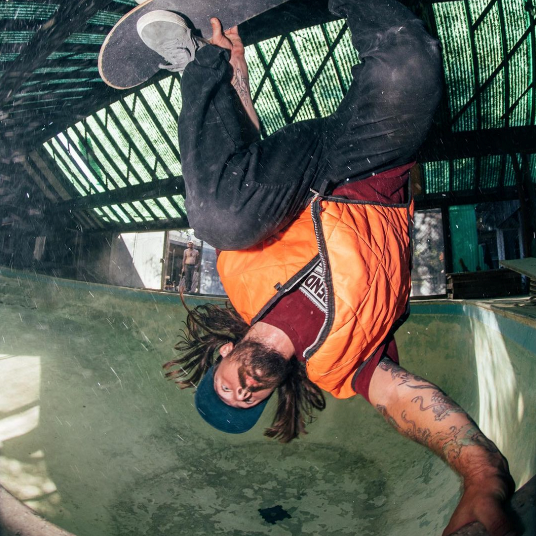 Kevin Kowalski doing a handplant in an empty pool, wearing an orange vest and teal cap
