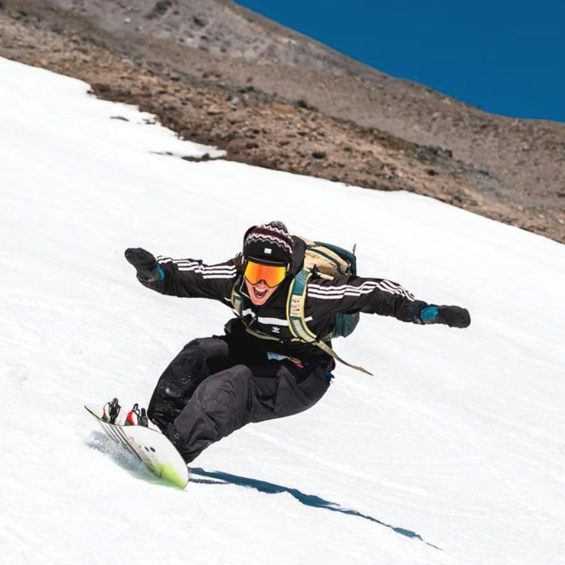 Luke Lund carving a snowboard down a steep snowfield in orange goggles and a black jacket