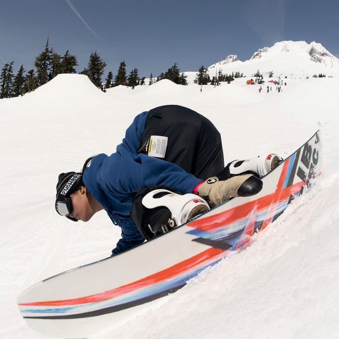 Nali Prevedel pressing a snowboard nose into the snow at a terrain park below a snowy peak