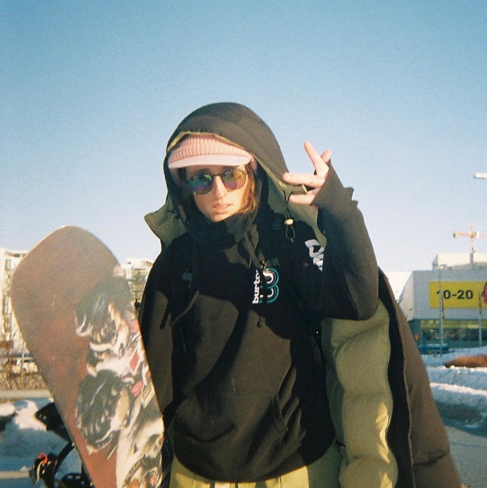 Nora Beck in sunglasses and a hooded puffer, holding a snowboard and flashing a peace sign