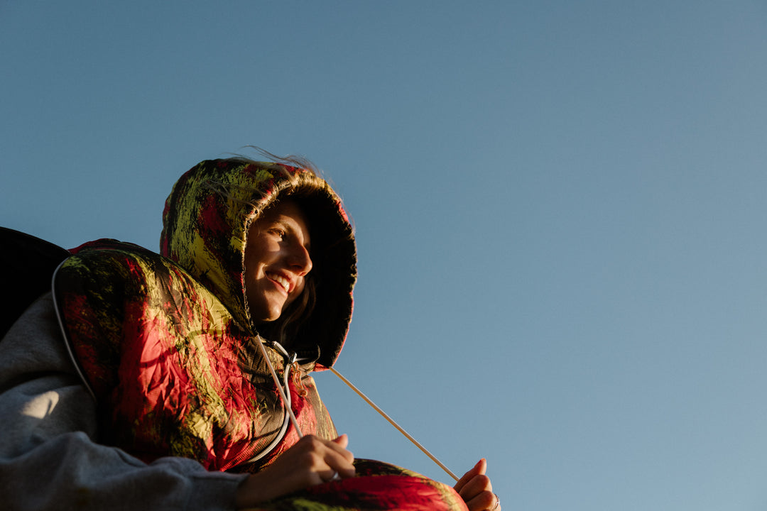 Smiling woman in the hood of a red and yellow print Napsack, lit by low sun against a blue sky