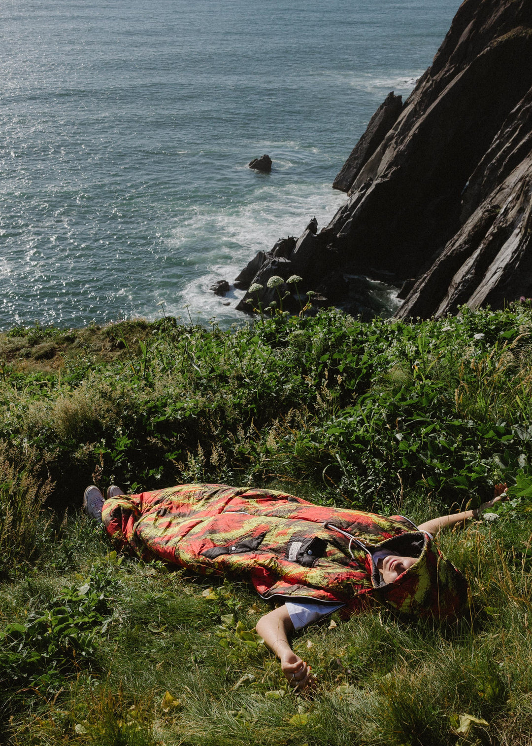Person lying in the grass on a seaside cliff wearing a red and yellow print Napsack
