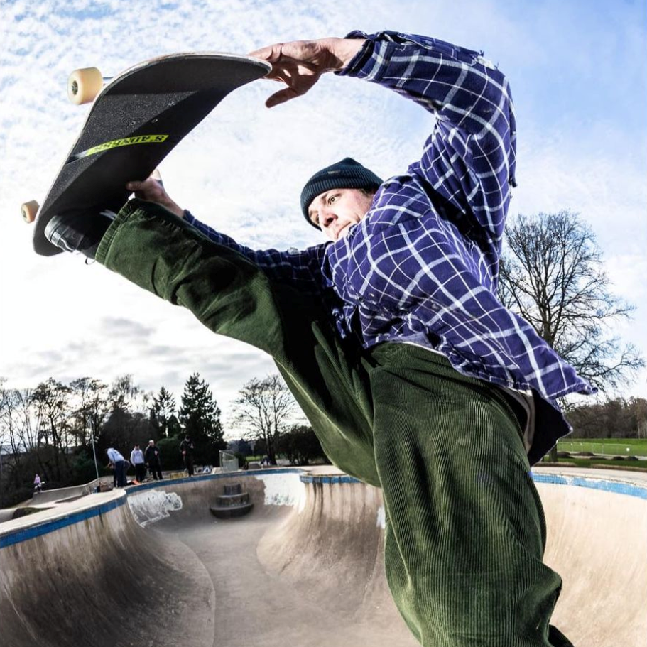 Ryan Price grabbing his skateboard above a concrete bowl in a blue plaid shirt and green cords