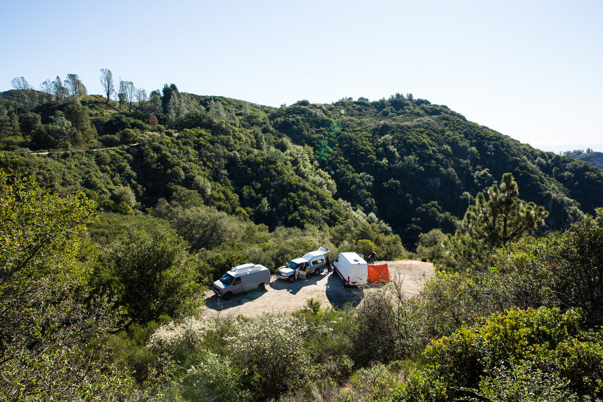 Vans and an orange tent parked at a clearing on a forested green hillside under a clear sky