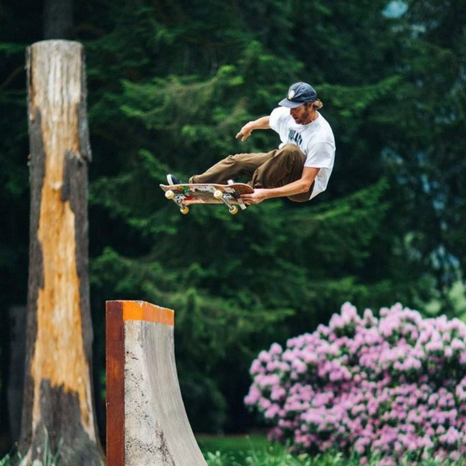 Willis Kimbel airing a skateboard off a wooden ramp beside pine trees and pink rhododendrons