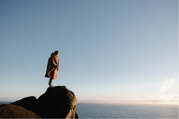 Person wrapped in a patterned Poler Blancho standing on a rock above the ocean at sunrise