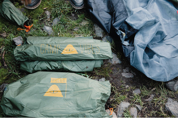Green Poler Camp Vibes stuff sacks on grass and rocks next to a blue tent