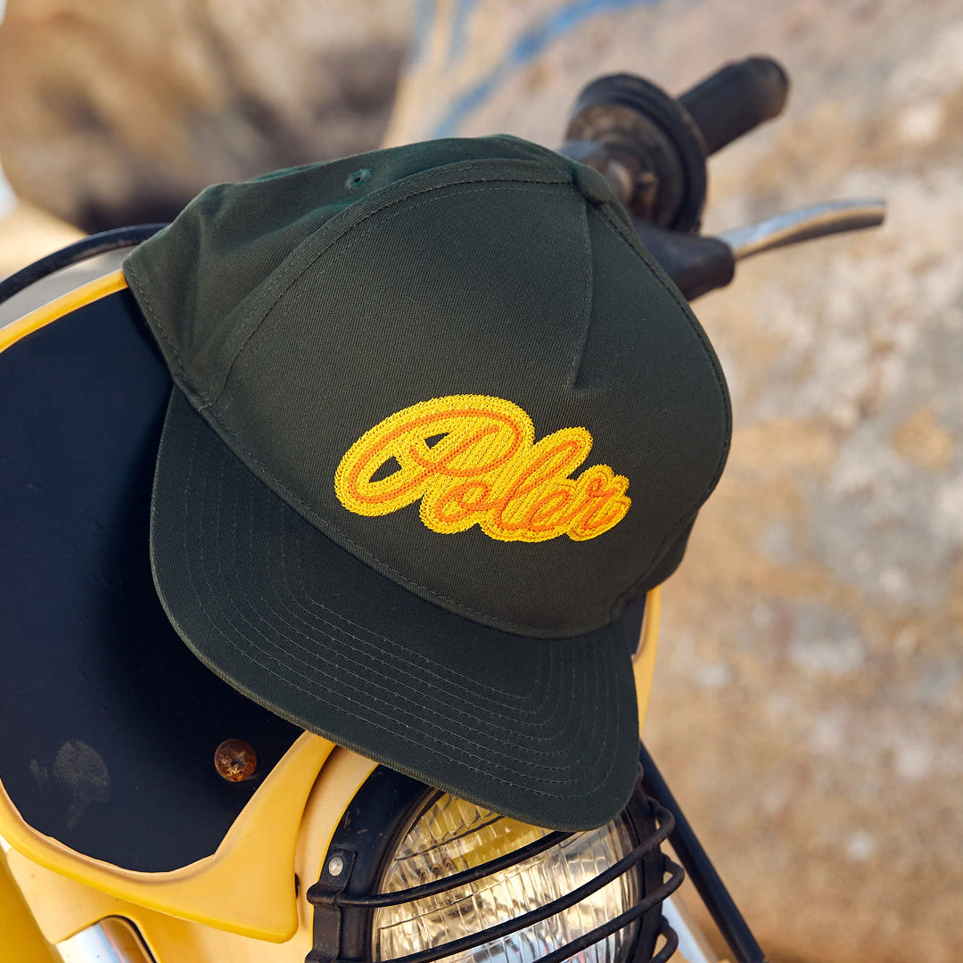 Dark green Poler snapback cap with orange chain-stitch script logo resting on a yellow motorcycle