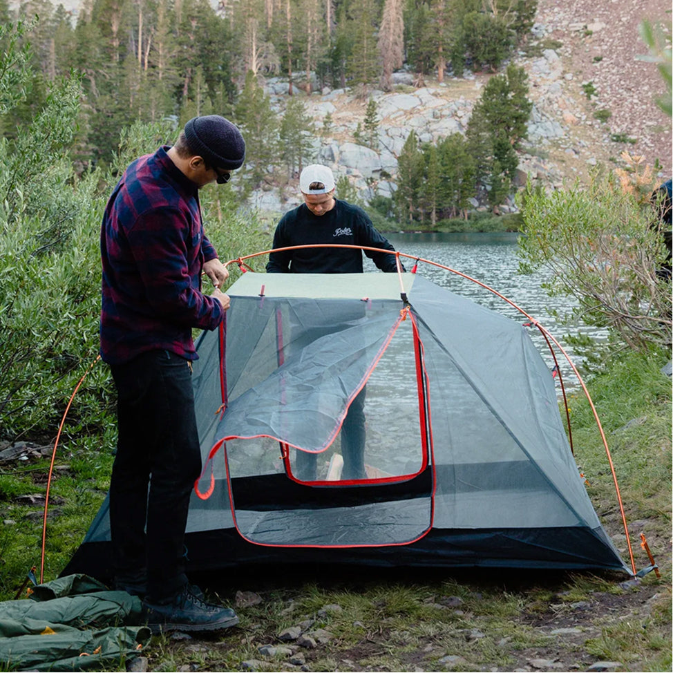 Two campers pitching a gray Poler tent with orange poles beside an alpine lake surrounded by pines