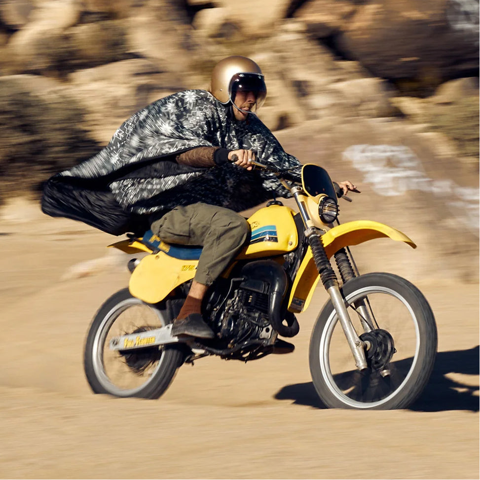 Rider on a yellow dirt bike kicking up dust in the desert, wearing a gold helmet and a patterned blanket poncho