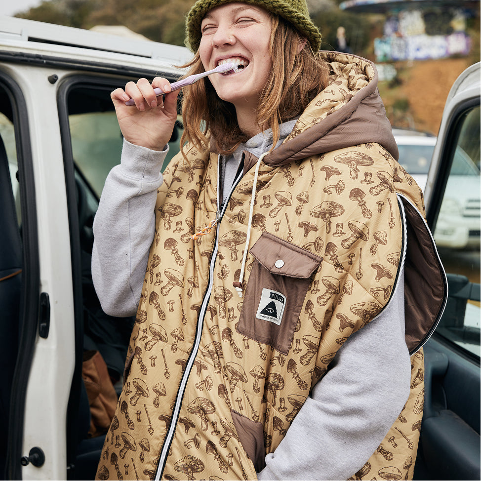 Smiling woman brushing her teeth by a van, wearing a tan mushroom-print Poler Napsack over a gray hoodie
