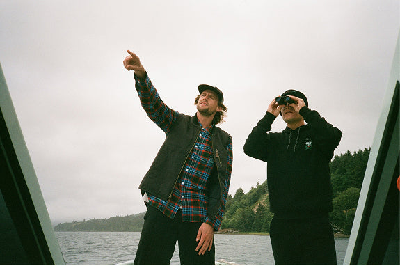 Two men on a boat, one in a flannel and vest pointing, the other in a black hoodie using binoculars