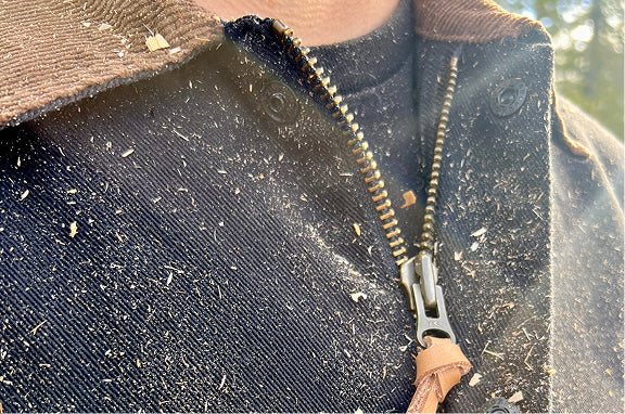 Close-up of a dark canvas work jacket with corduroy collar, brass zipper and leather pull, dusted with wood shavings