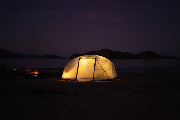 Poler dome tent glowing at night on a lakeshore beach beside a small campfire