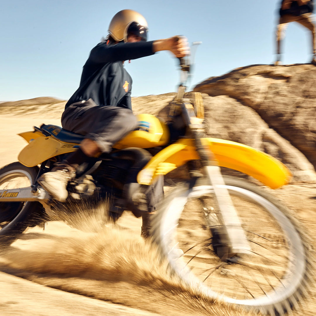 Rider on a yellow dirt bike spraying sand in the desert, wearing a gold helmet and dark jacket