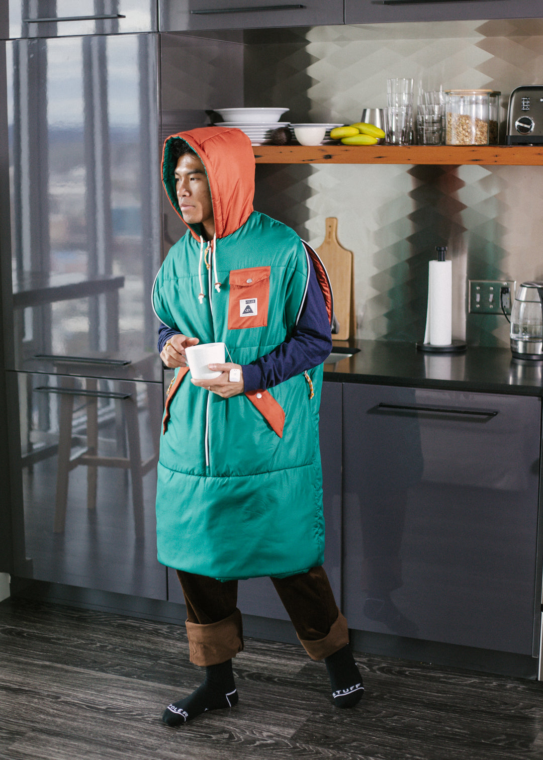 Man standing in a kitchen holding a mug, wearing a hooded teal and orange Napsack as a long vest