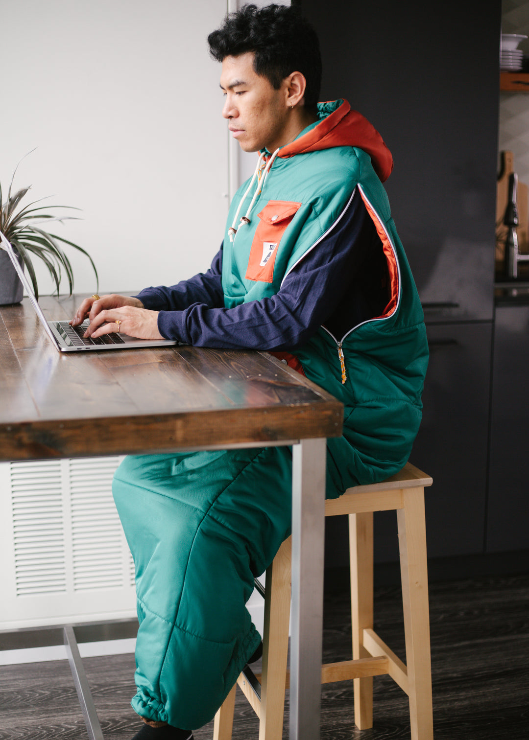 Man working on a laptop at a wooden table while wearing a teal and orange Napsack