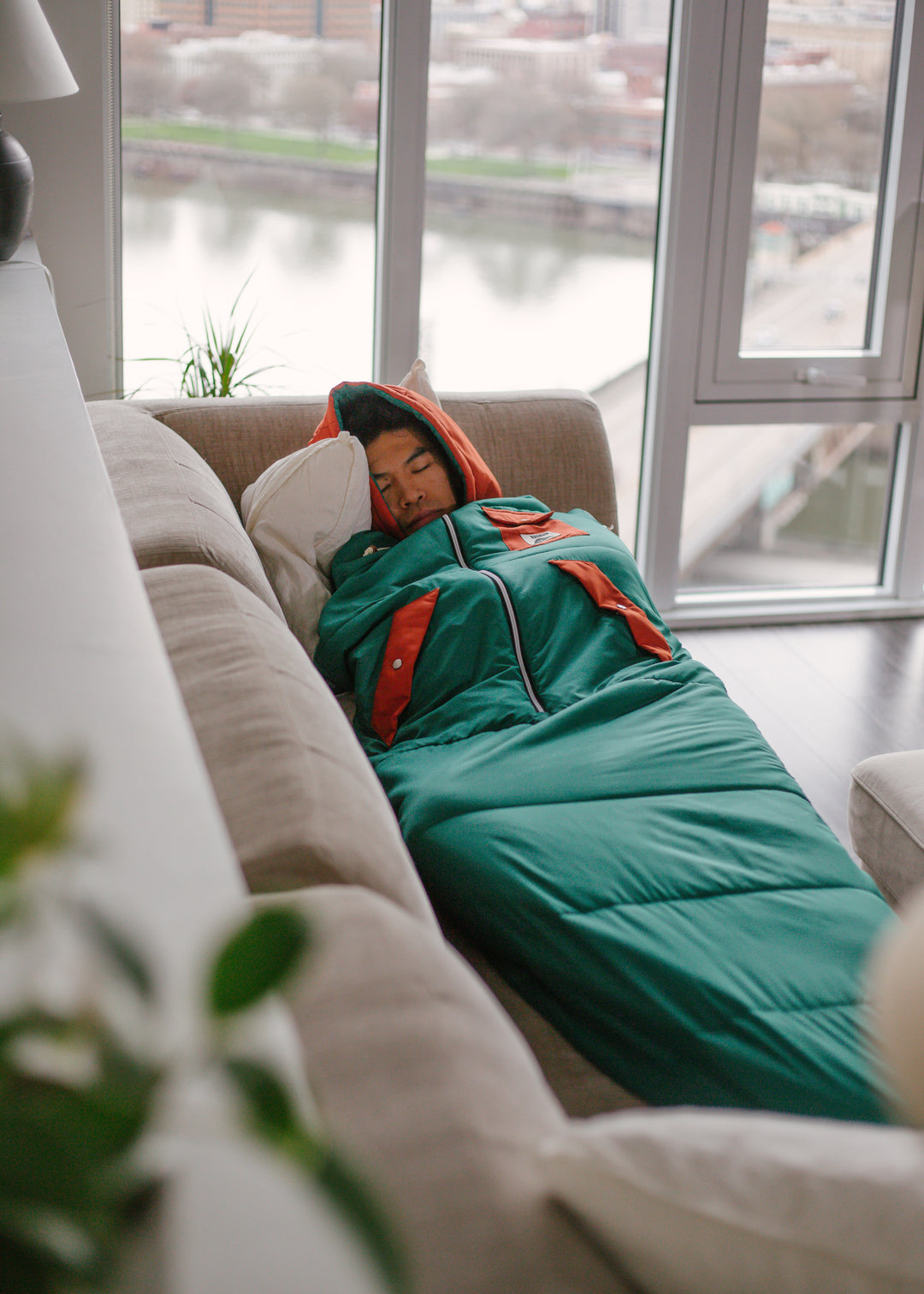Man napping on a couch zipped into a teal and orange Napsack, with a river view through the windows