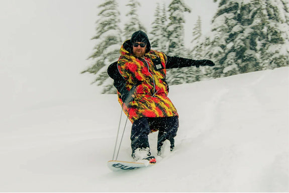 Snowboarder riding fresh powder wearing a red and yellow flame-print Poler Napsack