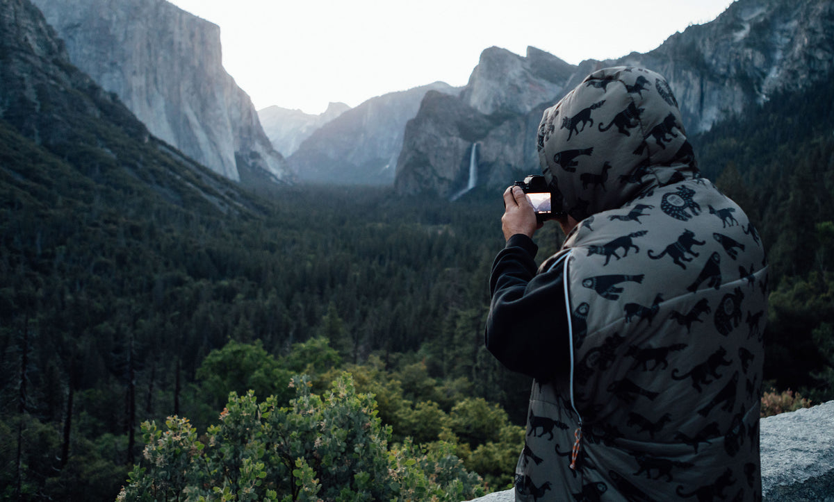 Person in a hooded animal-print Napsack photographing a valley of granite cliffs and a waterfall