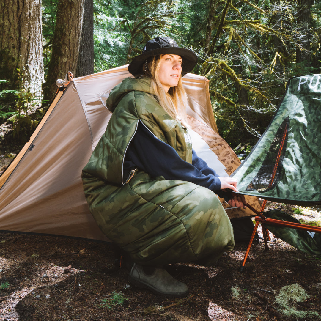 Woman in a camo Napsack and bucket hat crouching outside a tent in a mossy forest campsite