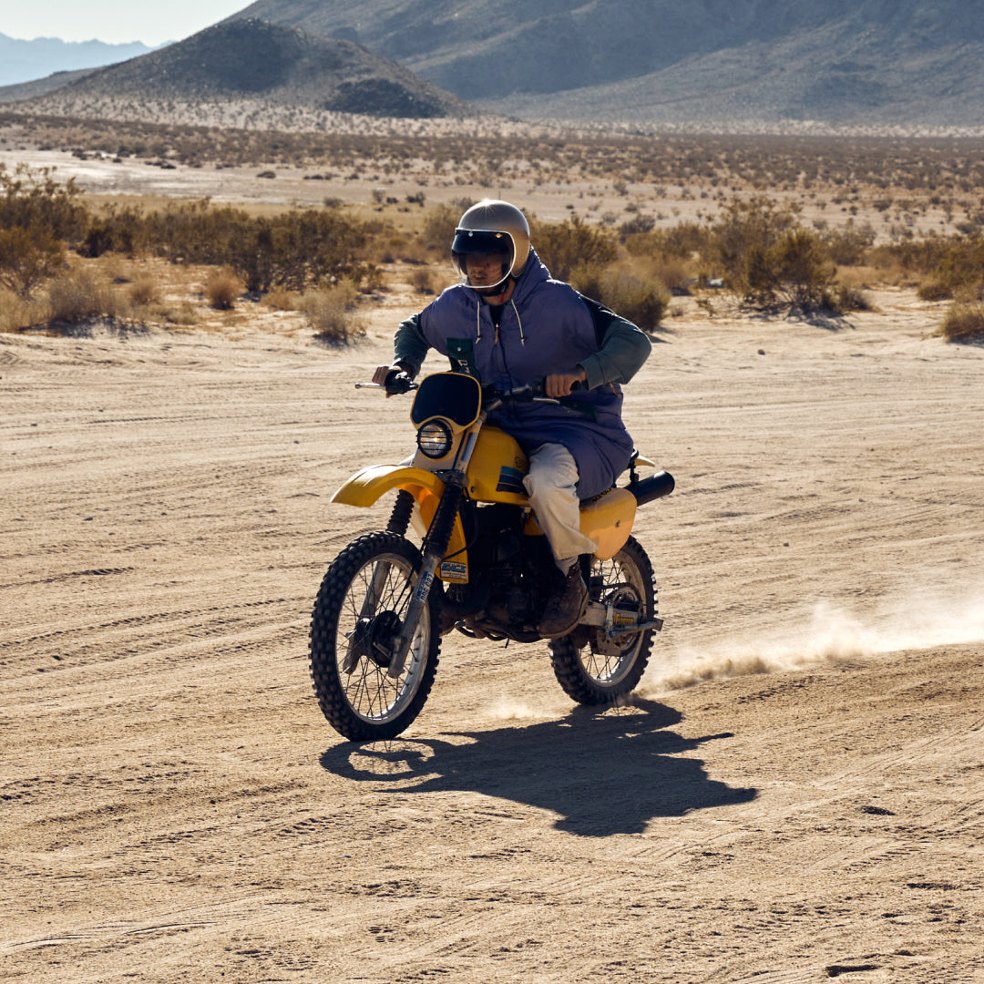 Rider wearing a purple Napsack on a yellow dirt bike crossing a sandy desert flat