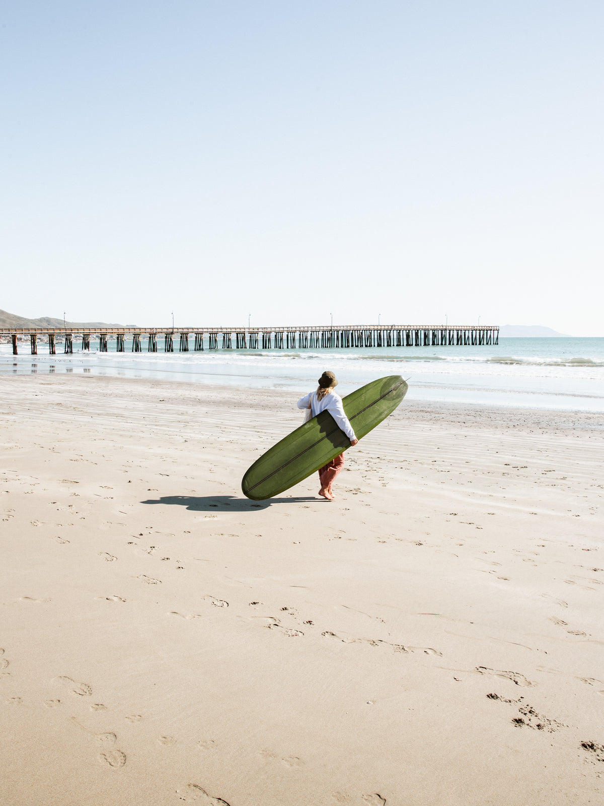 Surfer carrying an olive green longboard across a sandy beach toward a long wooden pier