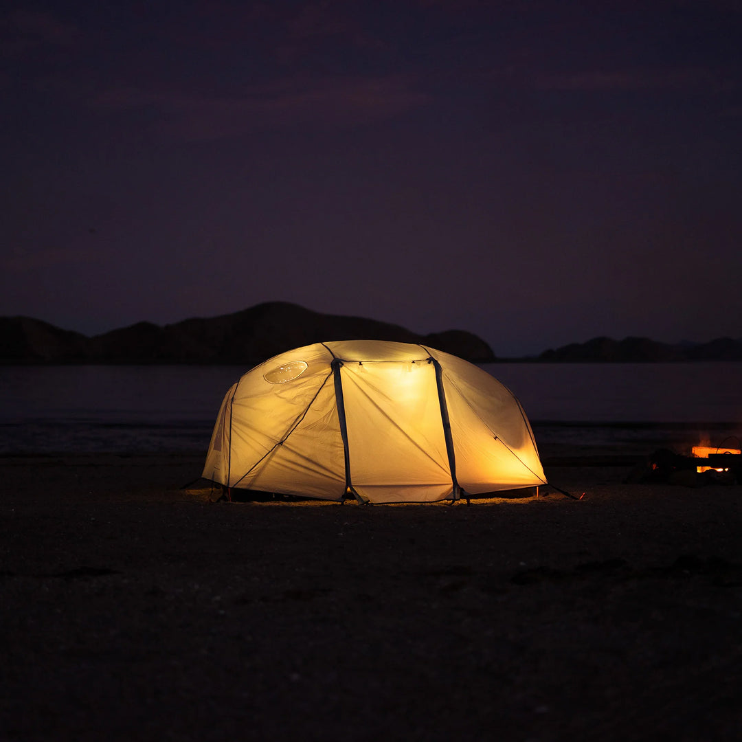 Dome tent glowing from inside at night on a beach, with dark hills and water behind