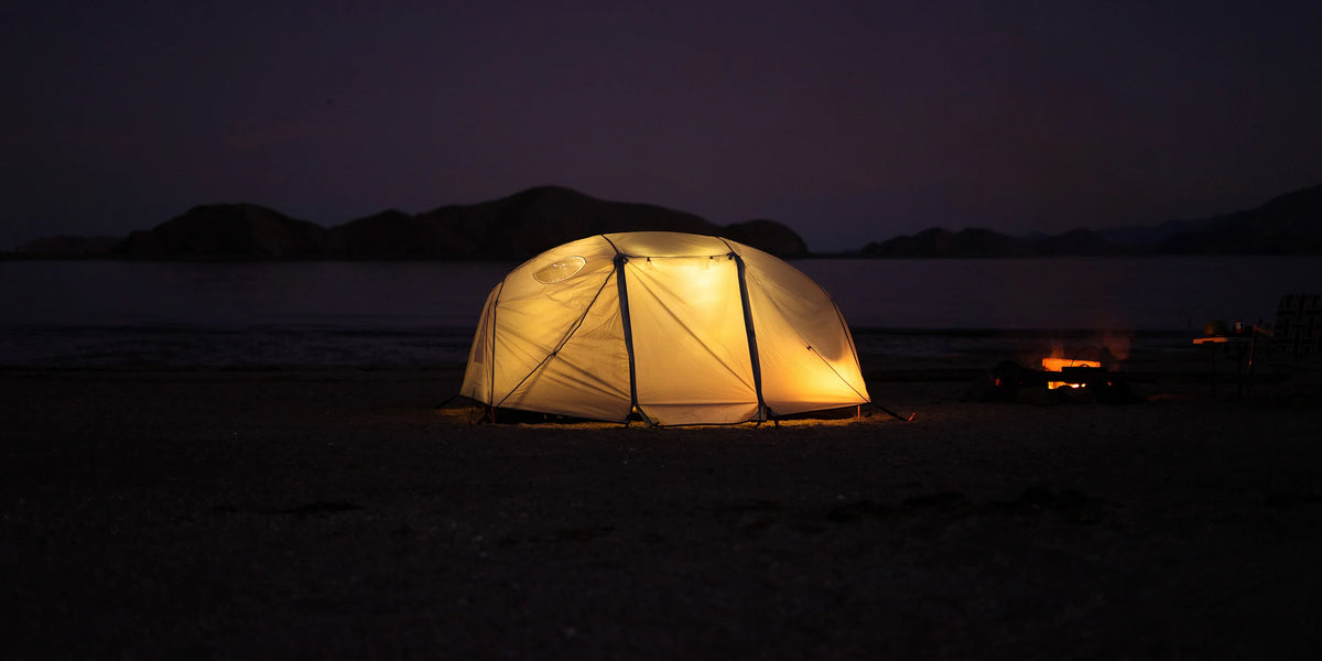 Dome tent glowing from inside on a beach at night beside a small campfire, with dark hills behind